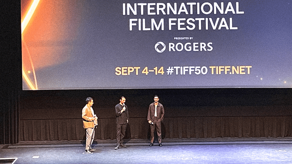 Three men with microphones stand on a stage in front of a screen with the general TIFF50 info slide.
