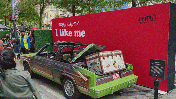 A burnt out 1983 Chrysler LeBaron with giant trunk full of stickers balancing atop its trunk. A red wall acts as backdrop with text: John Candy: I Like Me.
