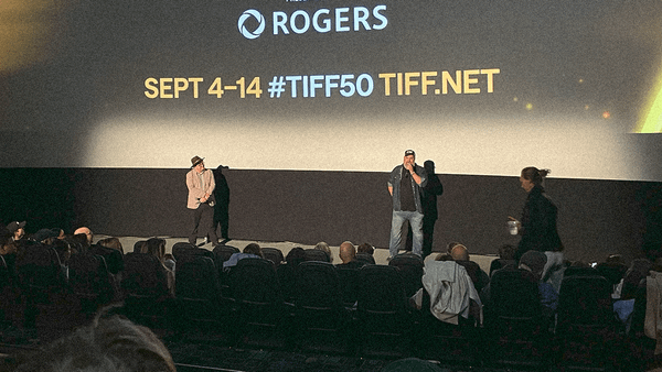 Two men stand before a movie theater audience with TIFF's branded info projected on screen behind them.