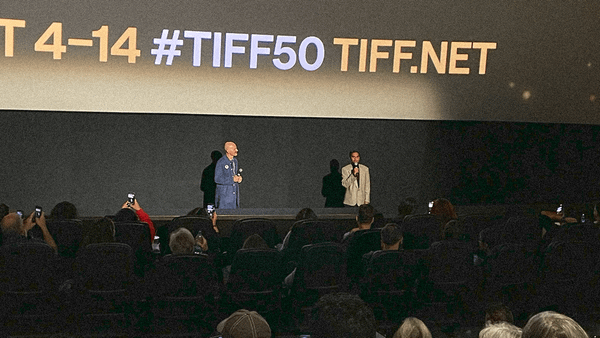 Two men with microphones standing in front of a full theater with the TIFF 50 brand info screen projected behind them.