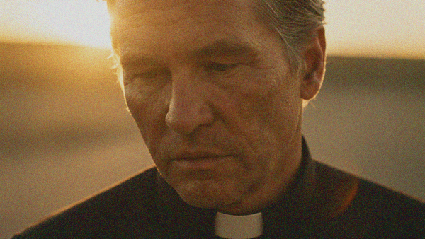 An aged priest in close-up looking downwards and forlorn as a bright sun shines behind him.