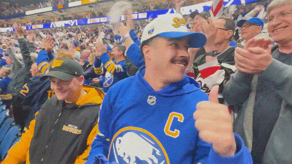 A man in Sabres apparel looks off-screen right with a smile and thumbs up while a man in Bruins apparel laughs to his right.