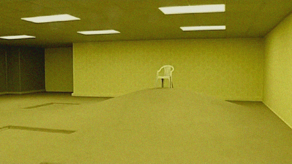 A strange, empty office-like room with a yellow tinge from wallpaper and lights where the floor rises into a mound and a white plastic chair stands atop it.