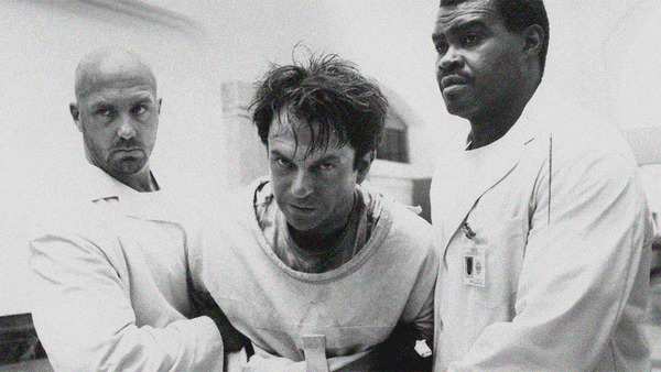 Sam Neill in straitjacket (flanked by two orderlies carrying him off-screen left) stares directly into the camera.