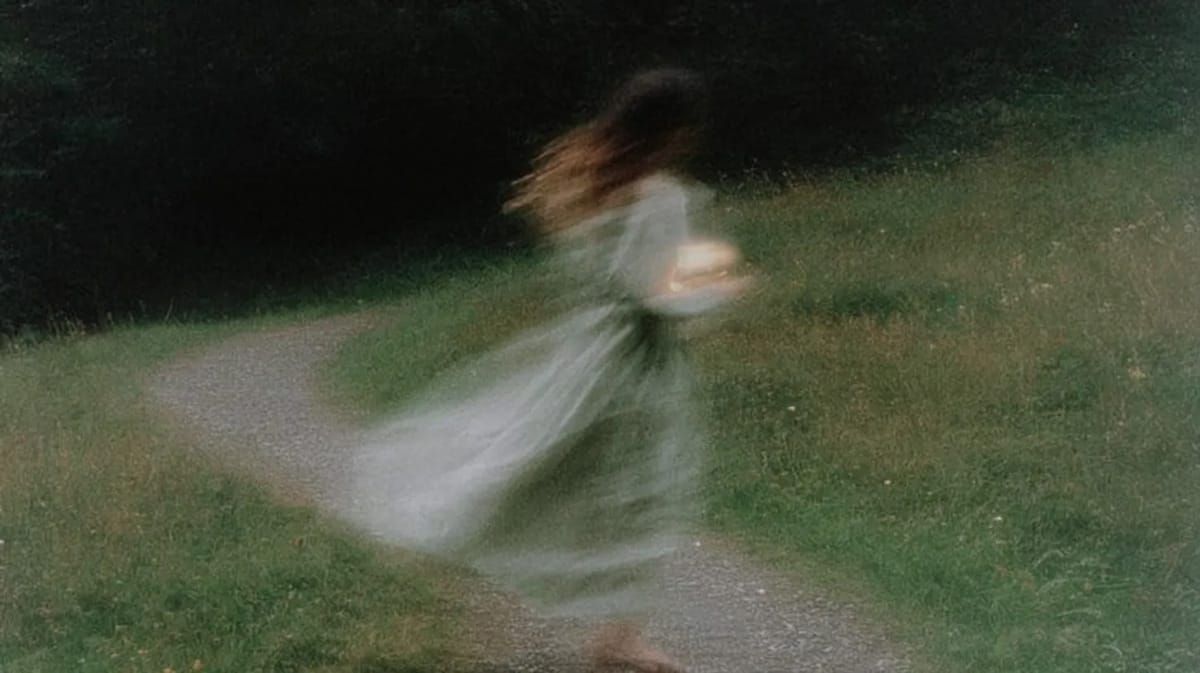 Soft-focus photo of a woman in a pale dress moving along a curving path in a green field, motion blur trailing her skirt. | Teachings from Khawaja Shamsuddin Azeemi.