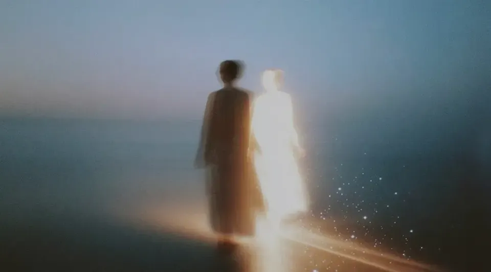 Soft-focus scene of a figure on a shoreline at dusk, one dark, one glowing white with sparkles, walking toward the horizon. | Teachings from Khawaja Shamsuddin Azeemi.