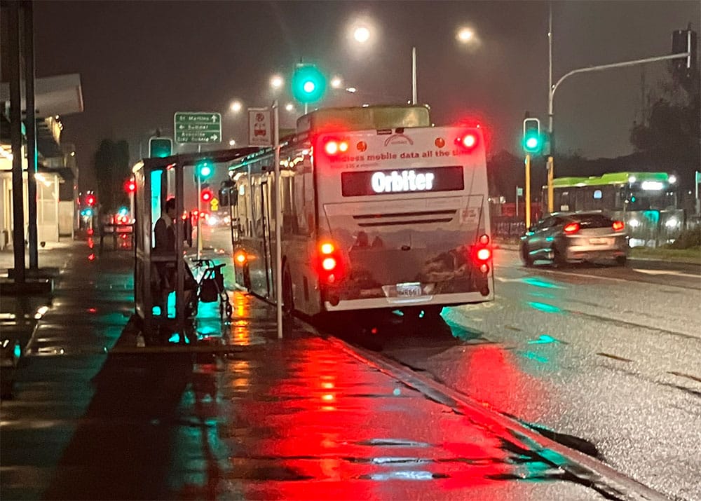 Photo taken at night looking at a bus from behind with its brake lights reflected on the wet road and footpath.