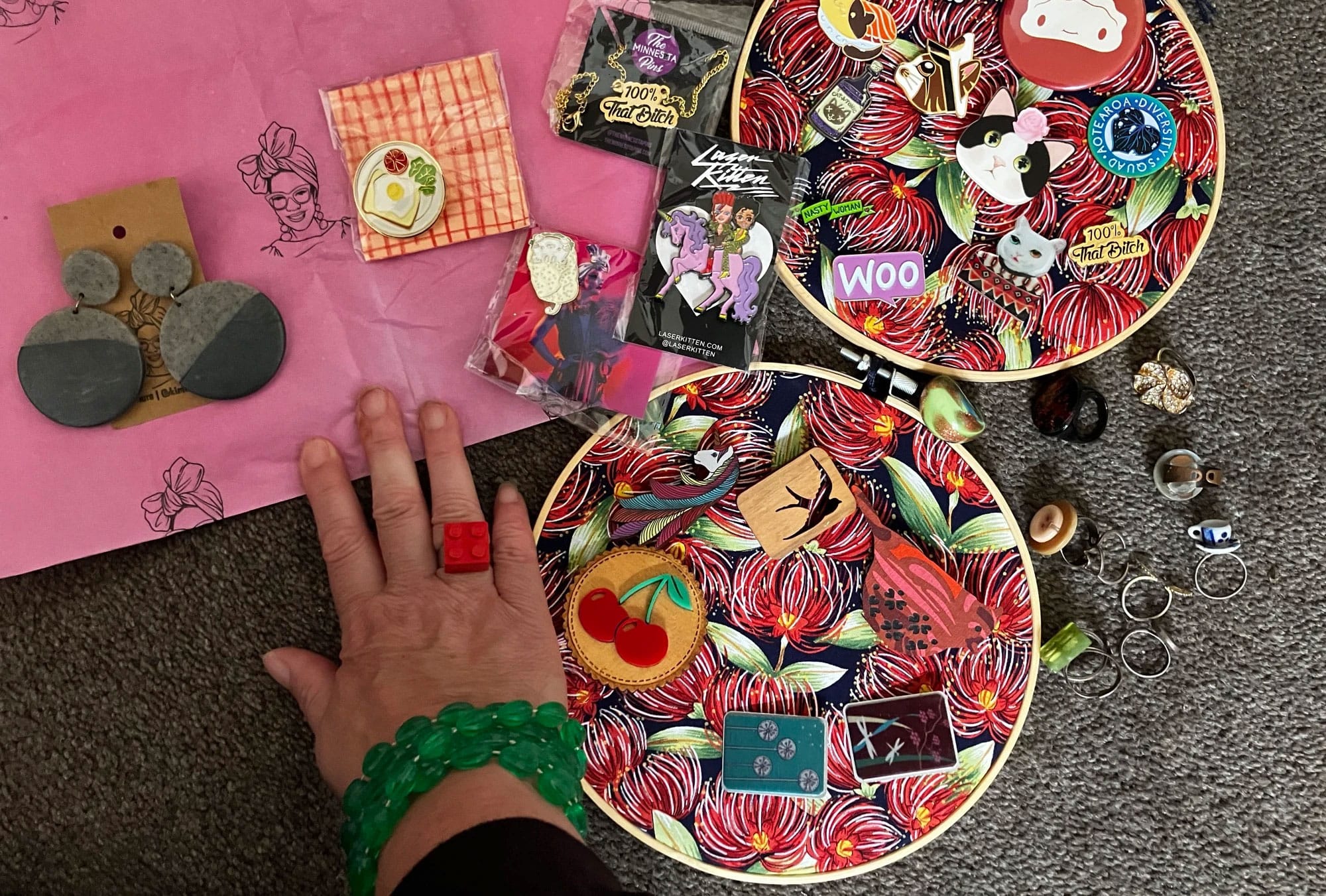 A couple of embroidery hoops with badges and pins attached to them, an assortment of rings and a pair of earrings made by Kim Tairi, the wahine, the legend.