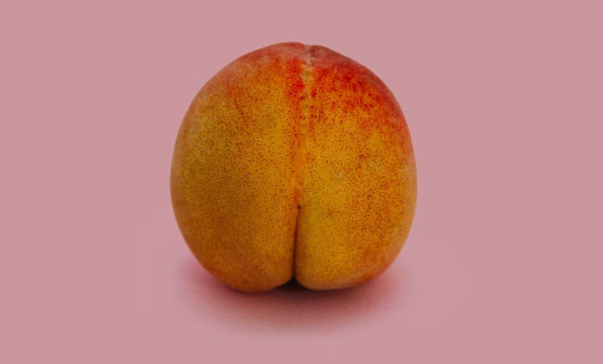 Photograph of a peach on a pink background. The peach does look a lot like a bum though.