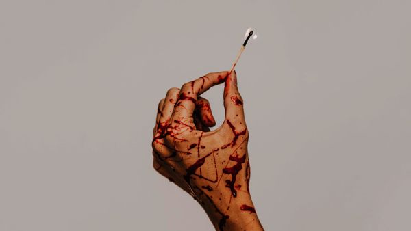 A woman's hand splattered with blood holds a lit match.