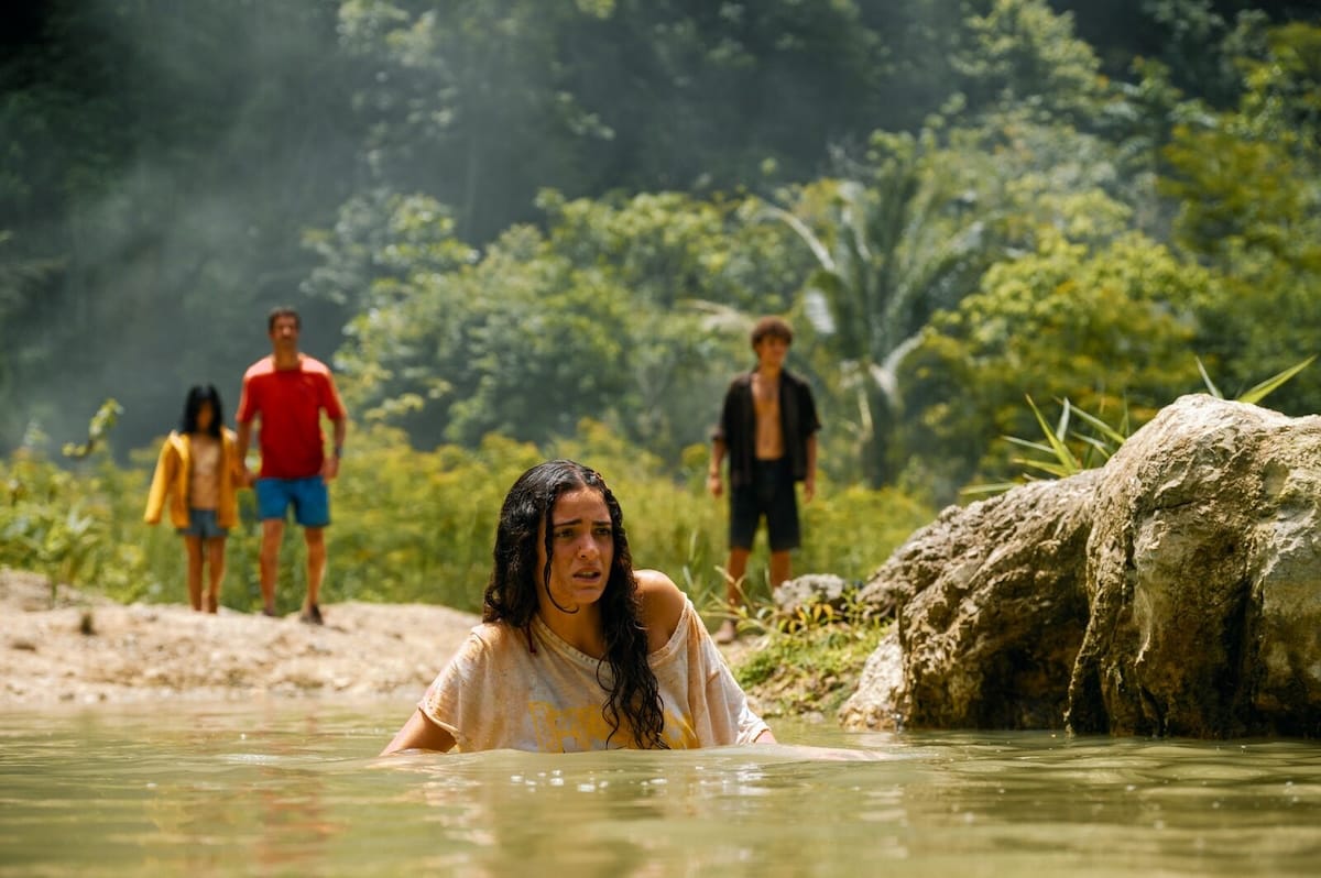 a young woman wades through a jungle river while her family watches worriedly from the shore