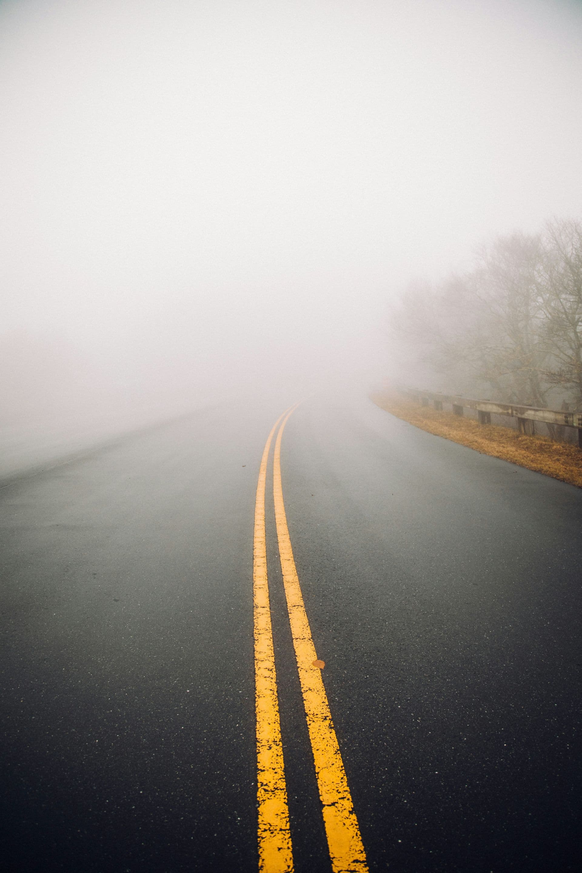 "A minimalist, high-contrast photograph looking down a long, wet asphalt road with two solid yellow lines disappearing into a thick, heavy white fog."