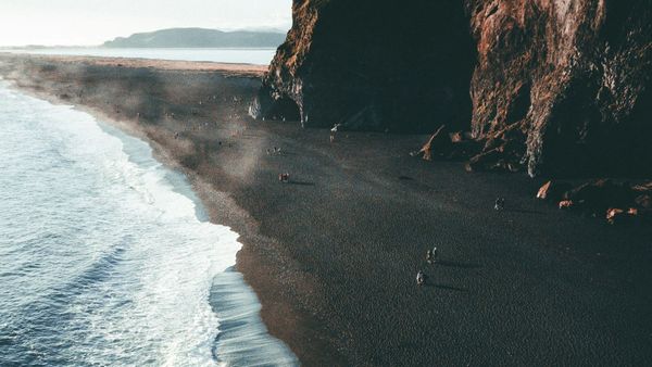 "A minimalist, high-contrast overhead photograph of a dark volcanic sand beach bordered by steep rugged cliffs, meeting the foam of white ocean waves."