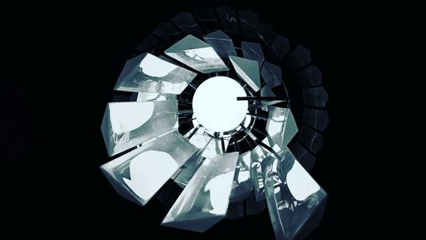 "Minimalist, high-contrast overhead photograph of a fractured geometric light fixture with metallic shards surrounding a bright central ring of light."
