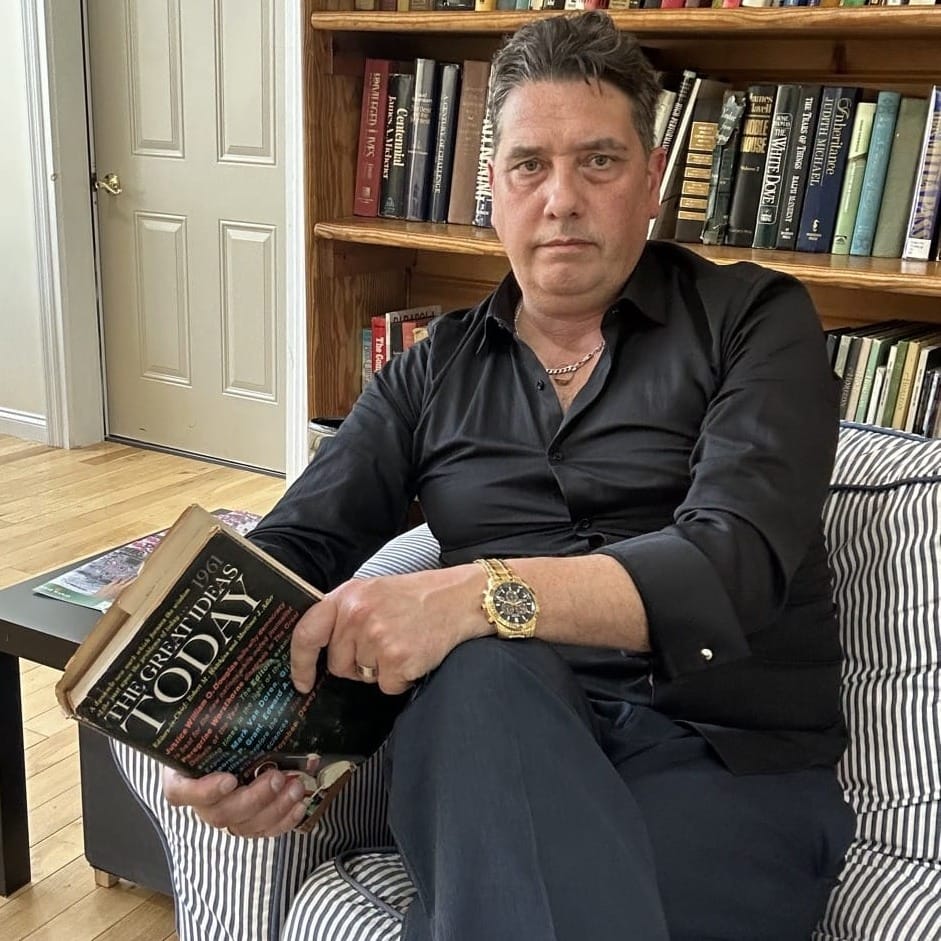 Elliott King, Editor of BEYONDai, seated in a striped armchair in front of full bookshelves, holding a hardback book.