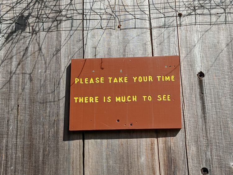 A painted wooden sign, yellow on brown, reading "PLEASE TAKE YOUR TIME THERE IS MUCH TO SEE"