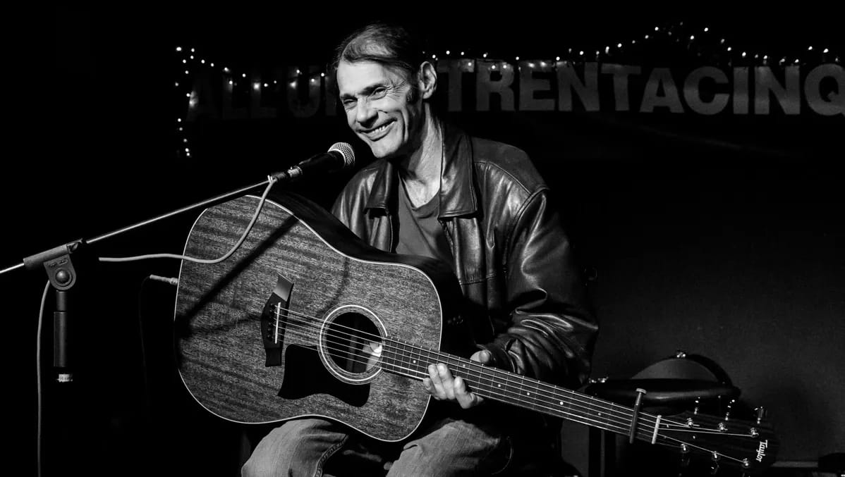Malcolm Holcombe sits on stage with his guitar, smiling at the crowd.