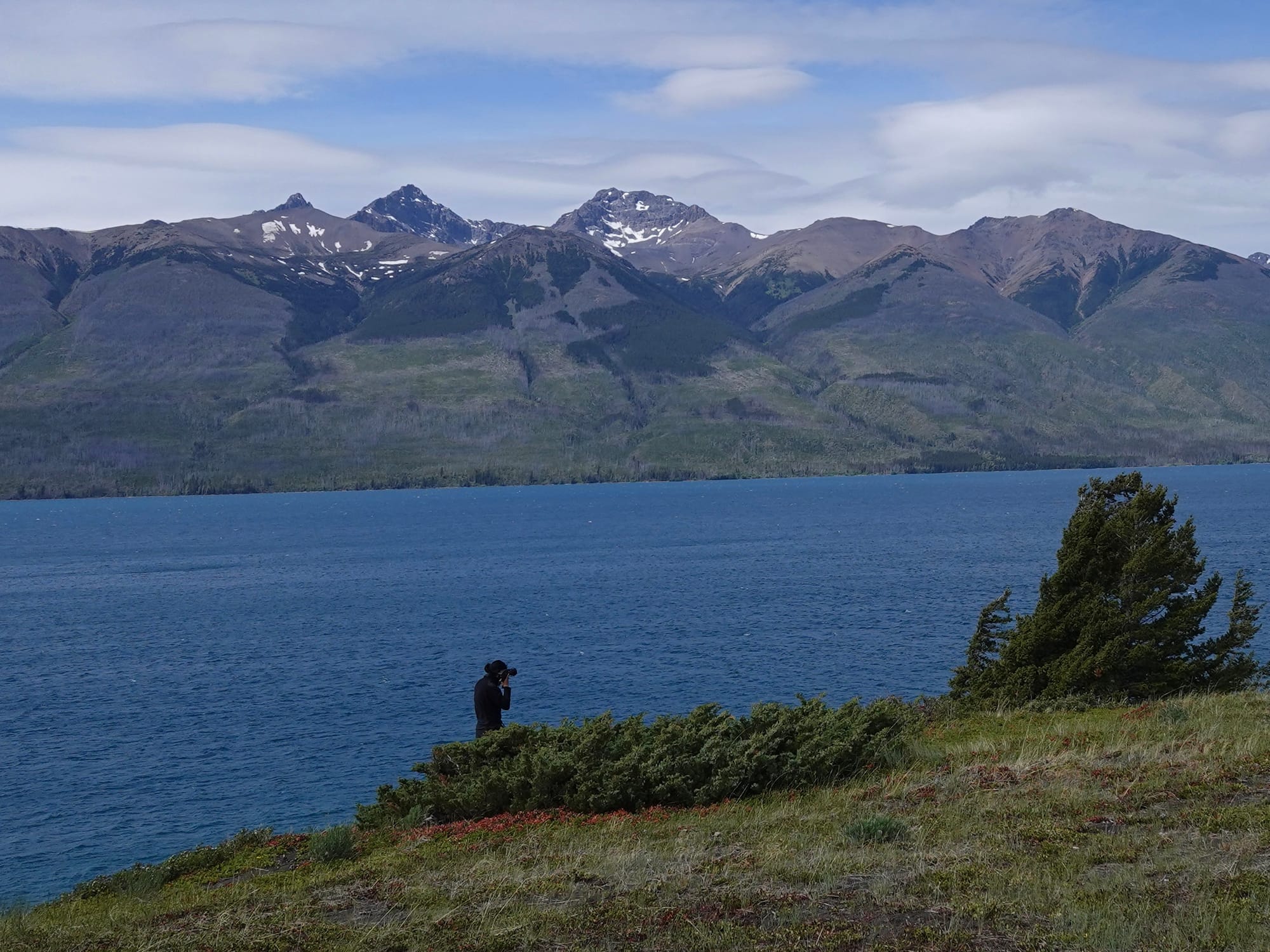 Shen Xin holding a camera to their ear standing between land and ocean, with snow topped mountains and blue sky afar