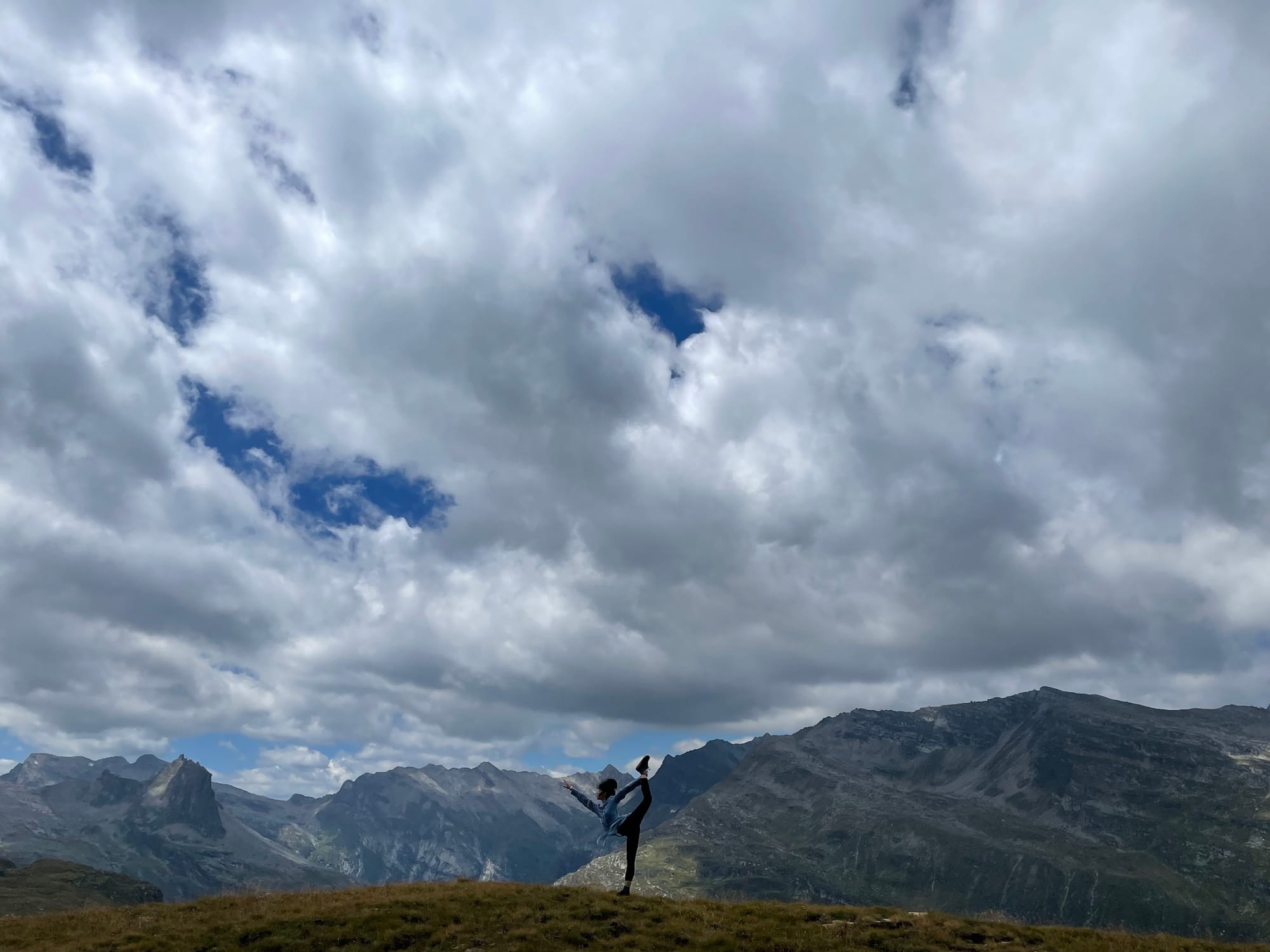 Ali lifting one leg up wearing blue shirt and black pants against mountainous landscape and large area of clouds