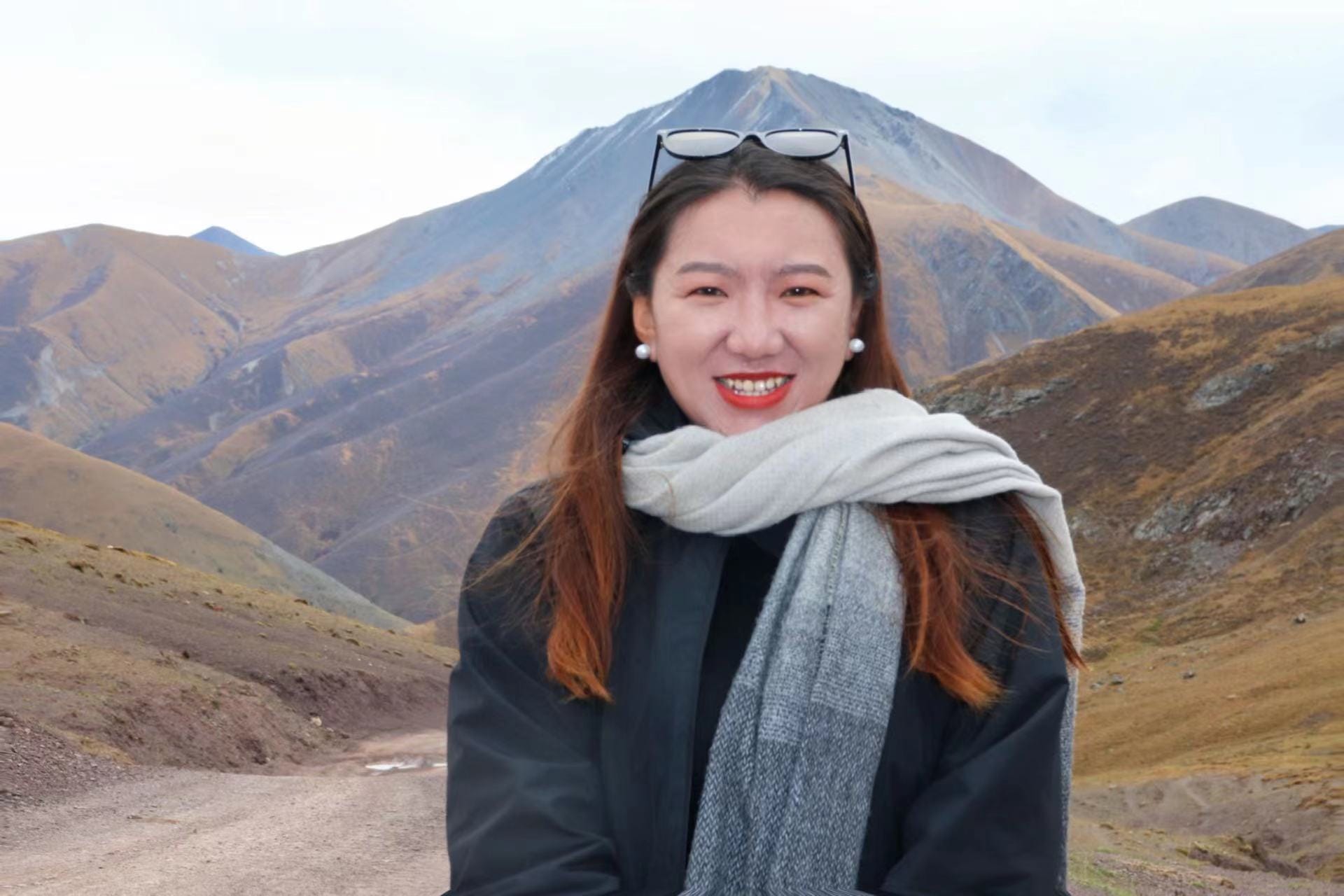 Skyid is smiling, standing in front of her mountain, wearing a white and grey scarf, with long orange coloured hair