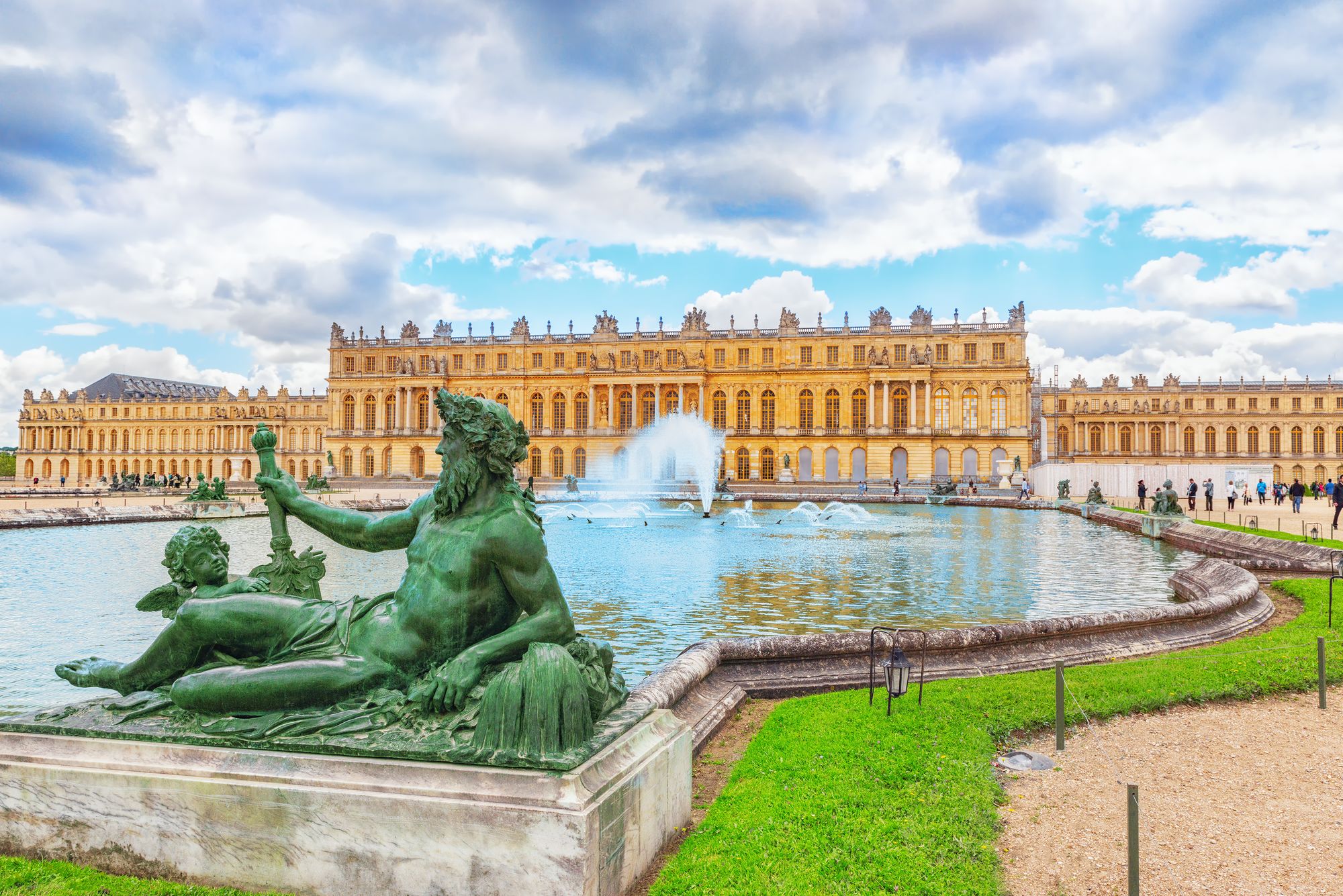 Una statua sovrasta la fontana nei giardini di Versailles, mentre in secondo piano domina la facciata della Villa