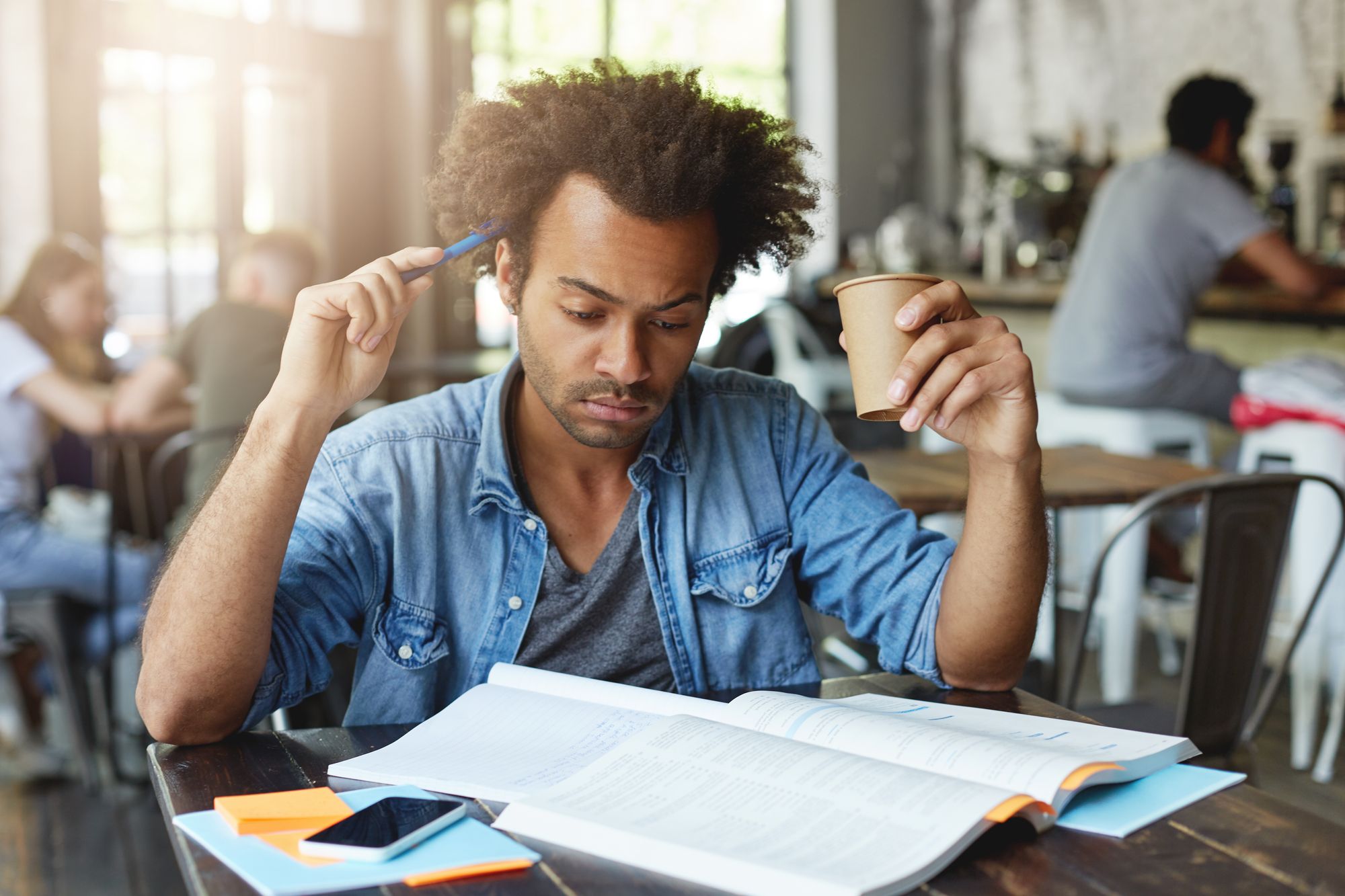 Concentrated thoughtful student with afro hairstyle scratching head with pen, drinking hot tea at cafe, preparing for lesson 