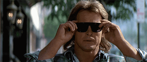 An animated GIF from 'They Live'. Nada (Roddy Piper) putting on sunglasses and seeing the hidden signs in the world including billboards saying consume