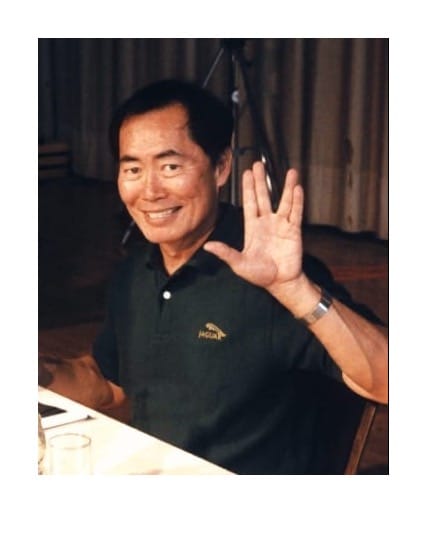“George Takei speaking into a microphone at a fan convention, wearing a shirt and jacket with a Star Trek backdrop behind him.”