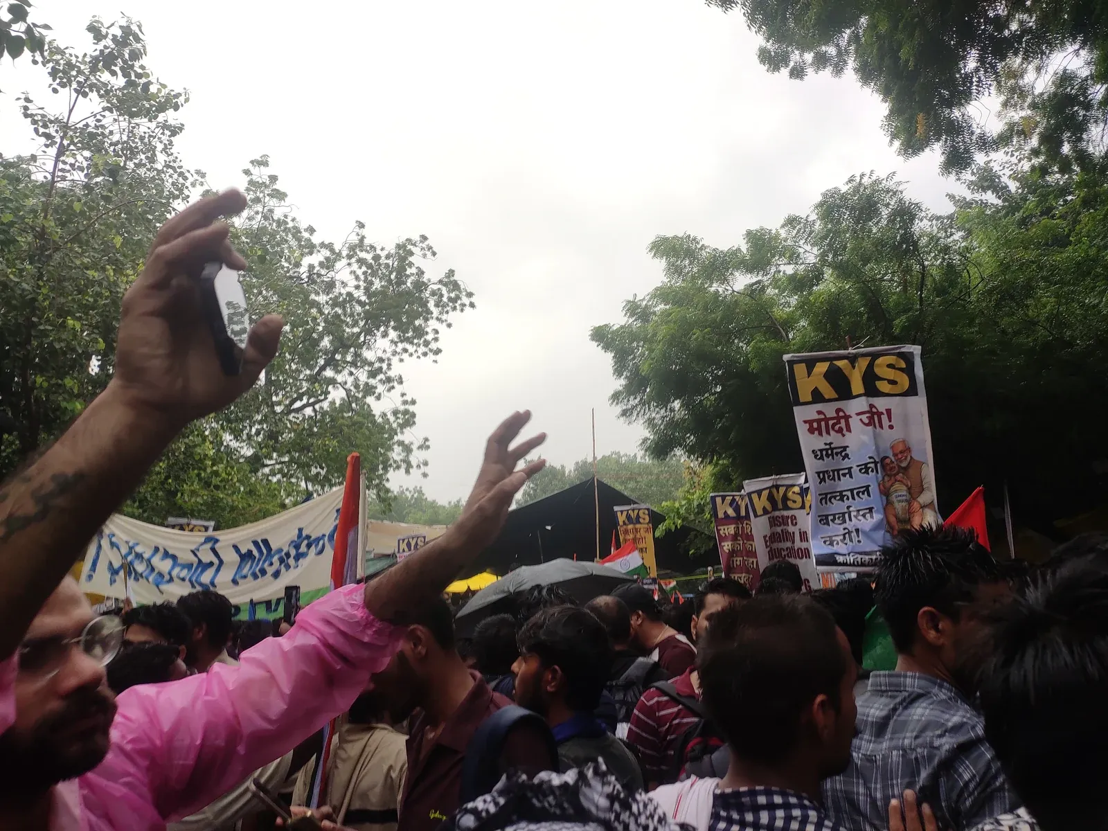 Protesters packed together in the rain at Jantar Mantar in New Delhi, umbrellas and banners overhead, one man in a pink shirt with his arm raised holding a phone above the crowd.