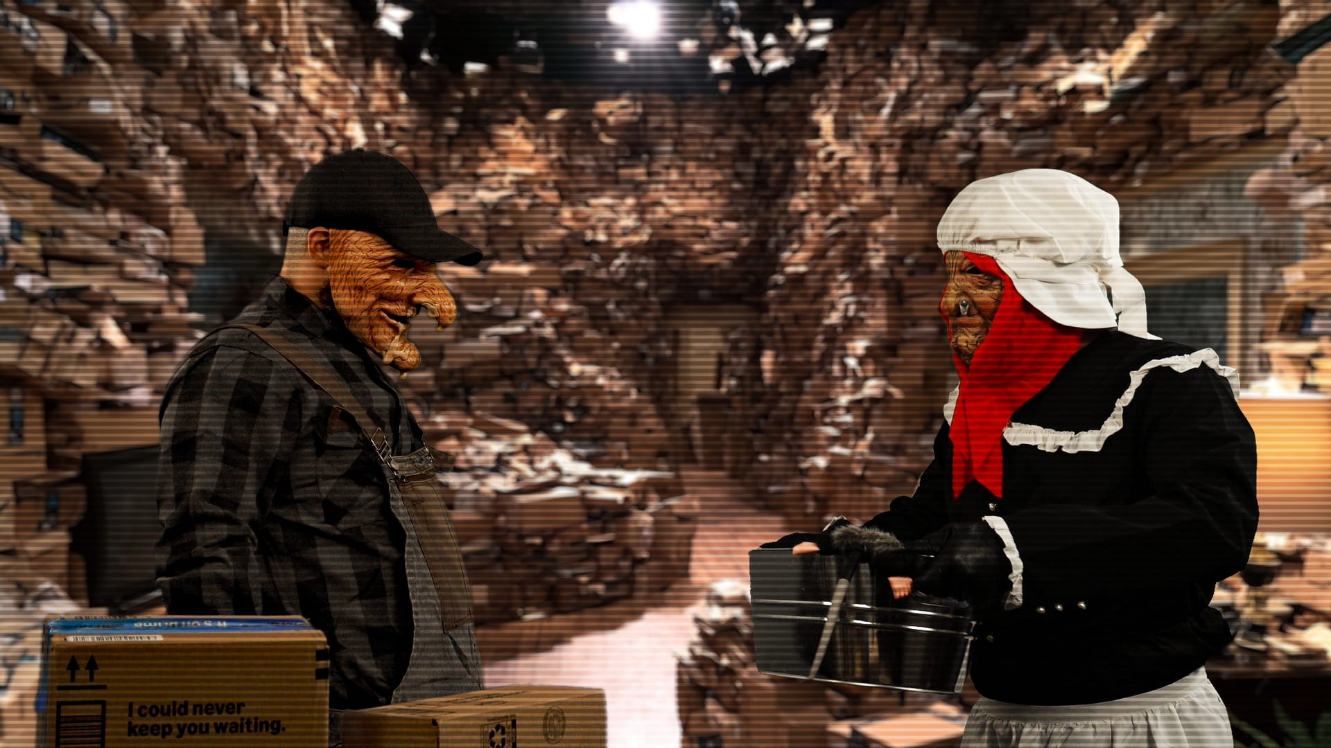 A masked performer in a black cap stands across from a masked performer in a white bonnet, red neck scarf, and apron inside a room built of stacked cardboard shipping boxes.