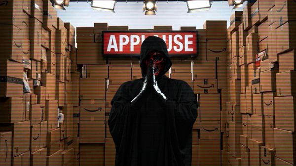 A red faced grim reaper stands under a red applause sign surrounded by Amazon boxes.