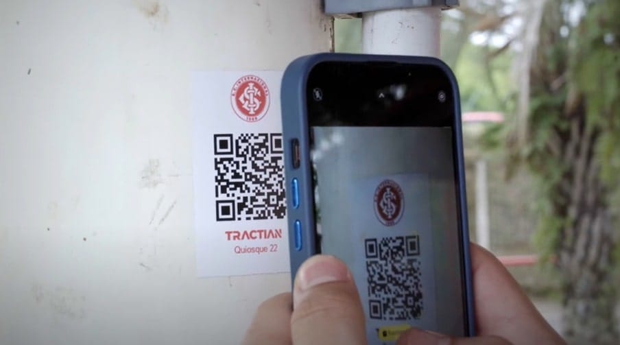 Um celular escaneia um código QR colado em uma parede. O adesivo contém o brasão do Sport Club Internacional. É possível ler TRACTIAN e Quiosque 22.