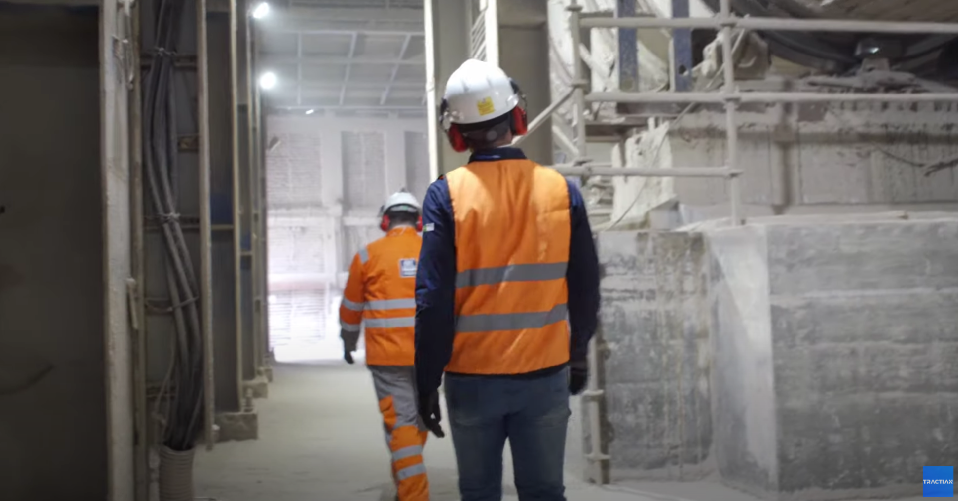 Dois homens andando em uma fábrica, vestindo coletes laranja e capacete.
