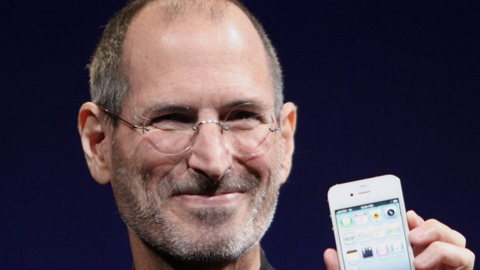 A picture of Steve Jobs, co-founder of Apple. He is holding an iPhone on stage at an Apple event.