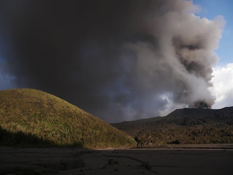 A gray ash cloud rising from the crater of Mount Semeru in East Java, Indonesia, with visible volcanic activity monitoring equipment in the foreground.