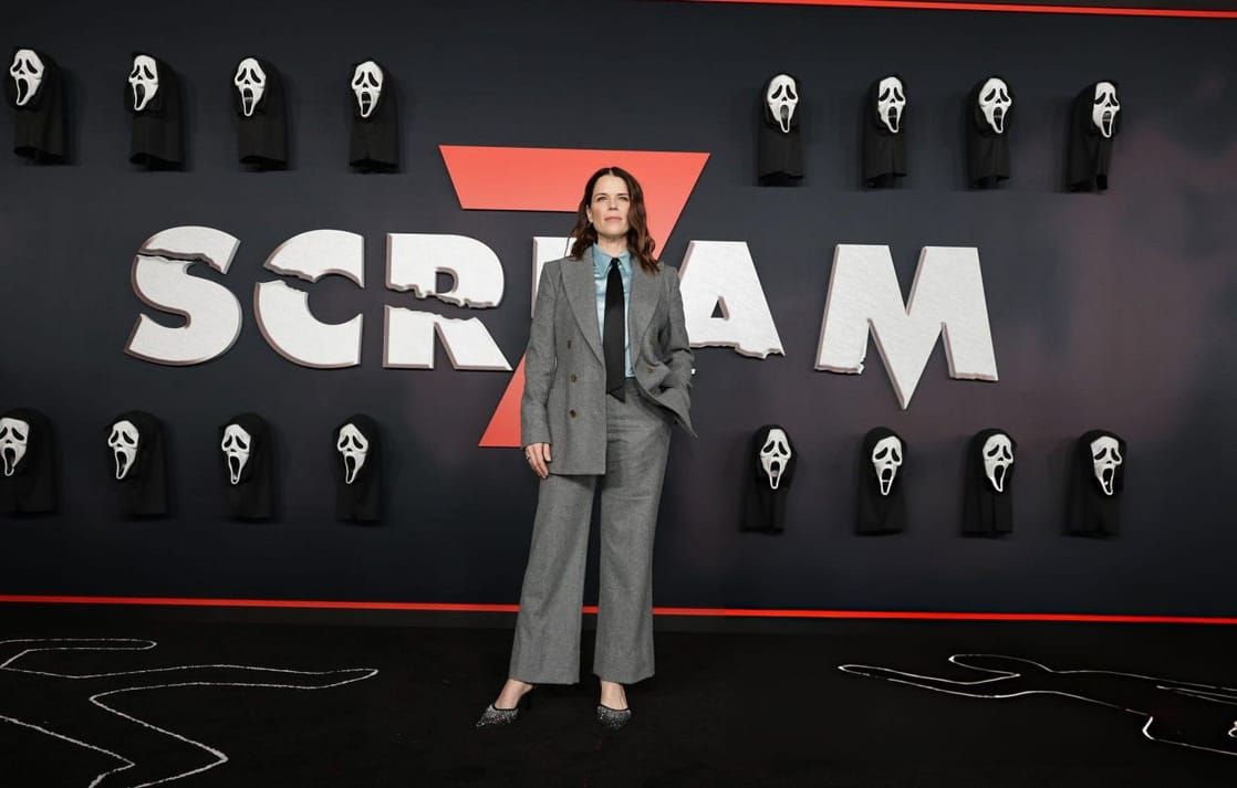  Actress Neve Campbell wearing a gray wool red carpet suit featuring a double-breasted longline blazer, wide-leg trousers, a blue satin blouse, and a black necktie at the 2026 Scream 7 premie