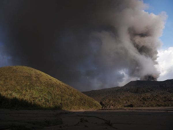 A gray ash cloud rising from the crater of Mount Semeru in East Java, Indonesia, with visible volcanic activity monitoring equipment in the foreground.