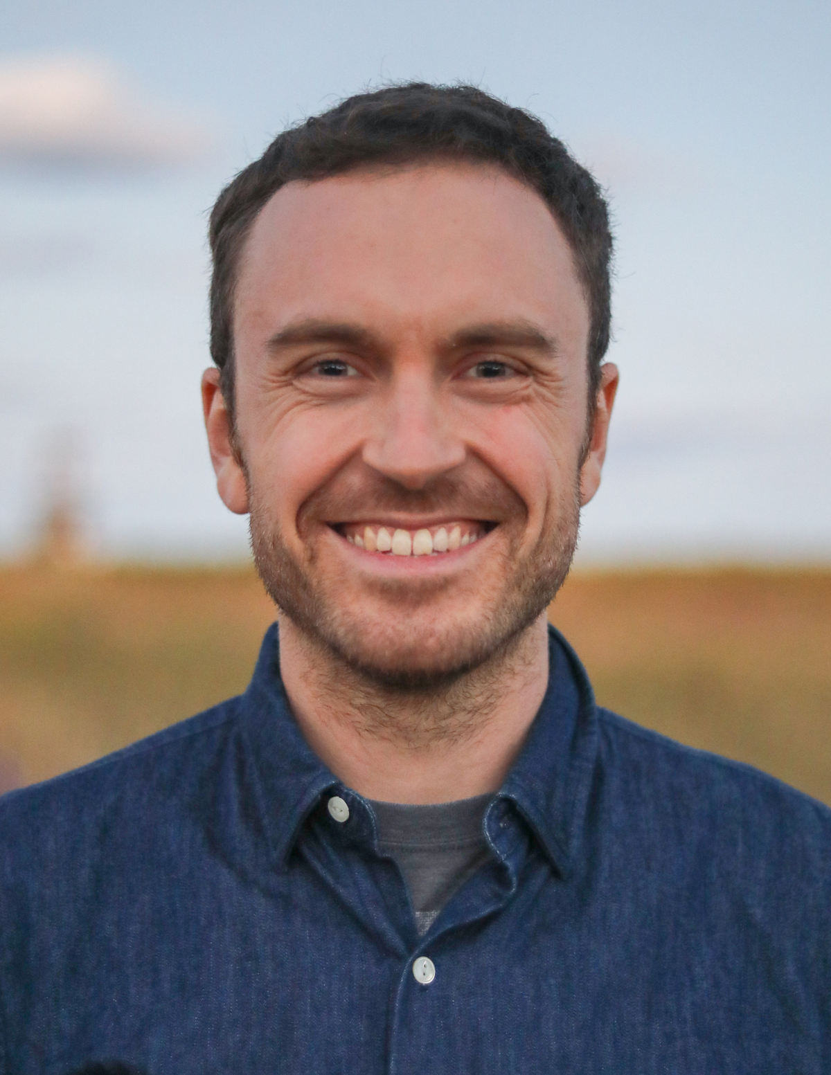 Smiling white man with brown hair and some facial hair, wearing a blue denim shirt with a grey t-shirt underneath, with a background of a yellowish brown blurred field background.