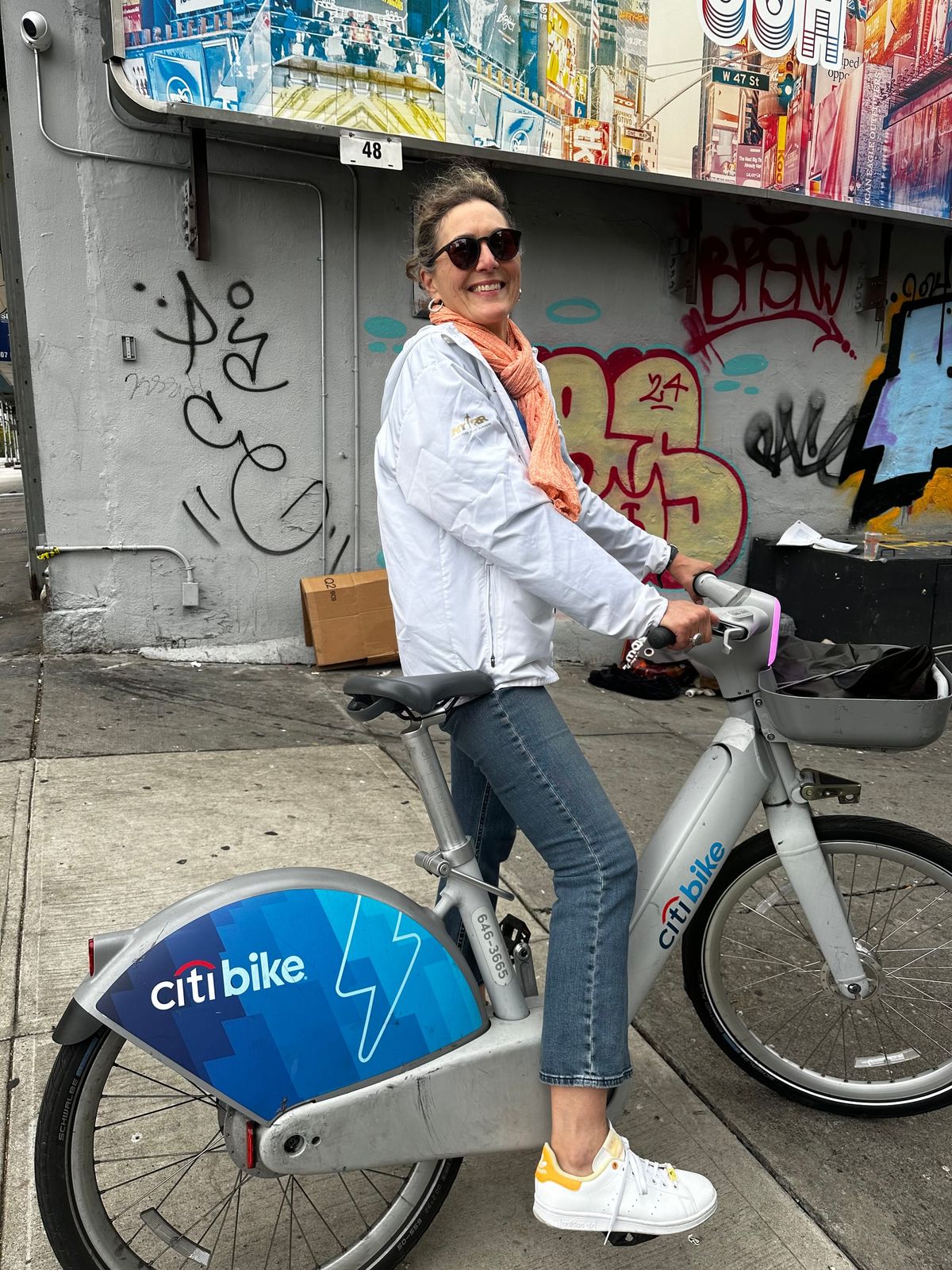 White woman with sunglasses in white rainjacket, jeans and white tennis shoes astride a Citibike e-bike on a NYC sidewalk next to a graffiti-covered wall. 