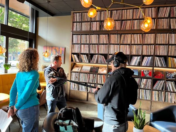 Three white people in foreground in a building with light coming in through window at left, facing a wall of vinyl records on shelves.  
