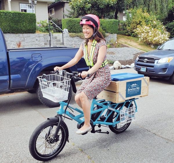 Asian woman with dark hair wearing a print dress with reflective vest riding a blue cargo ebike on a city street