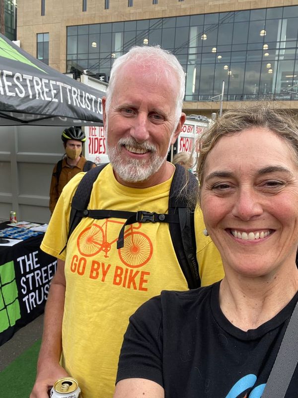 White man with facial hair in yellow shirt with "go by bike" on it and white woman with pulled back hair in black t-shirt alongside event tents
