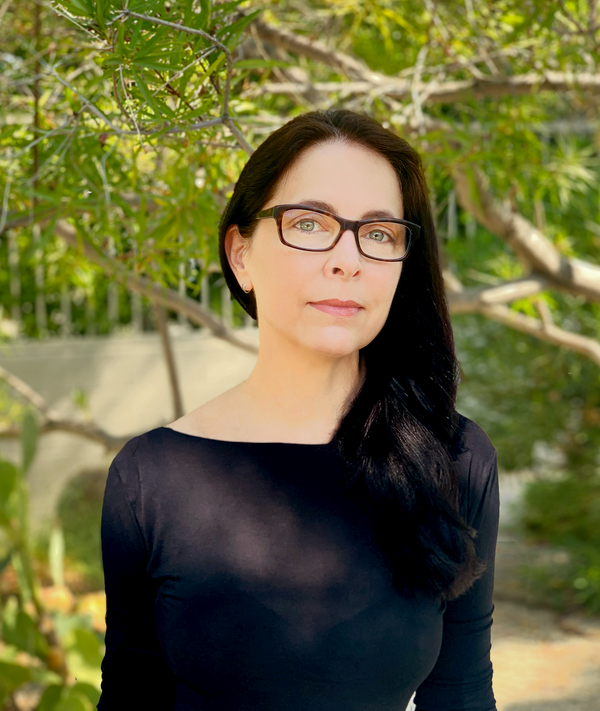 White woman with long straight dark hair and dark framed glasses in black, long-sleeved shirt sitting outdoors with some greenery and a patio wall behind her.  