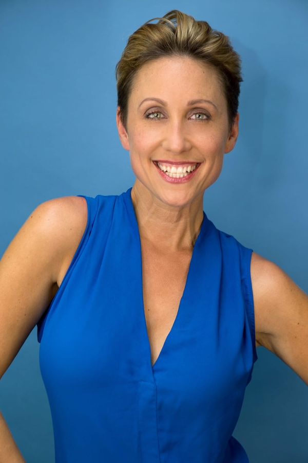 Smiling white woman with pulled back blonde hair, wearing a sleeveless blue dress.