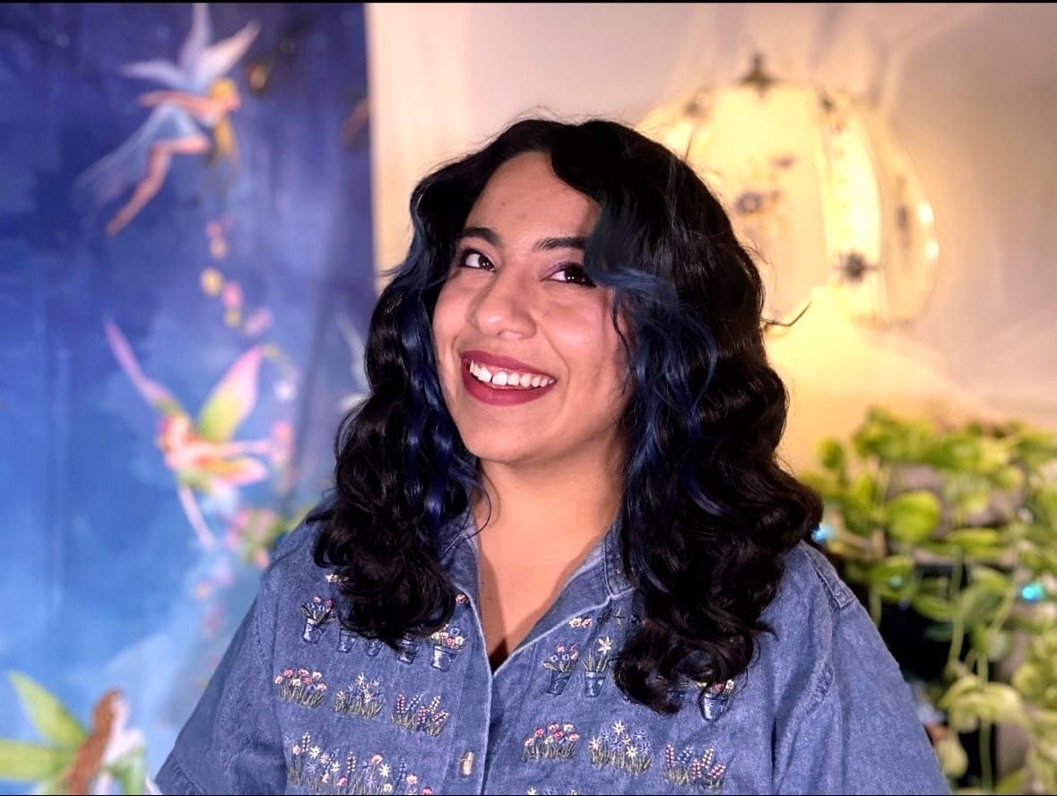 A photo of Winona Veil. She has brown wavy hair, a blue jean shirt with flower embroidery, and she has two blue highlights at the front of her hair.