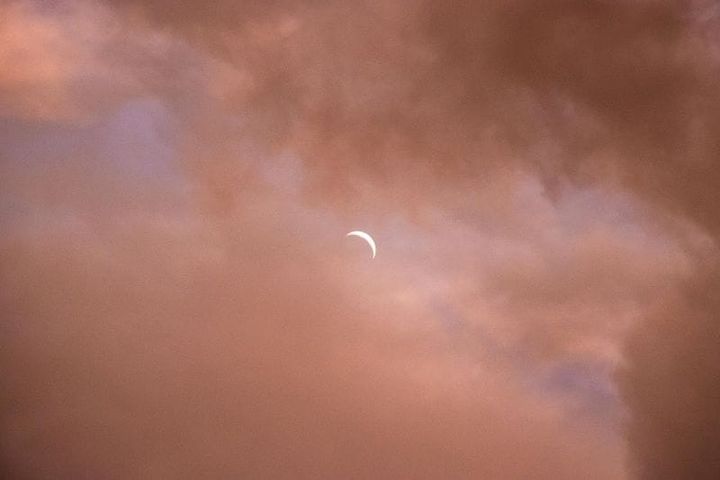 young waxing crescent moon, bright white against hazy pink clouds