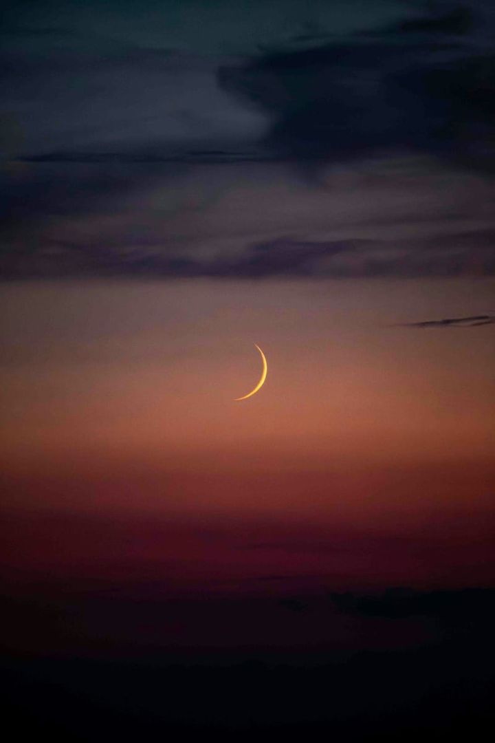 yellow sliver of a waxing crescent moon over sunset colours of peach, fuschia and deep purple