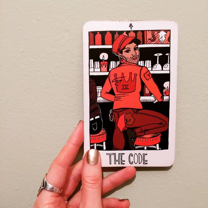 the code card from the collective tarot held up by a white femme's fingers with gold nail polish. the card shows a genderqueer aries leatherdaddy in red leathers at a bar. 