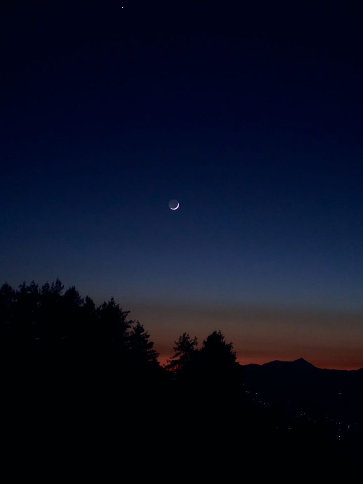 waning crescent moon, with the moon's circle visible by earthshine, in a clear sky at the end of a sunet. Silhouettes of evergreens and mountains in the foreground.