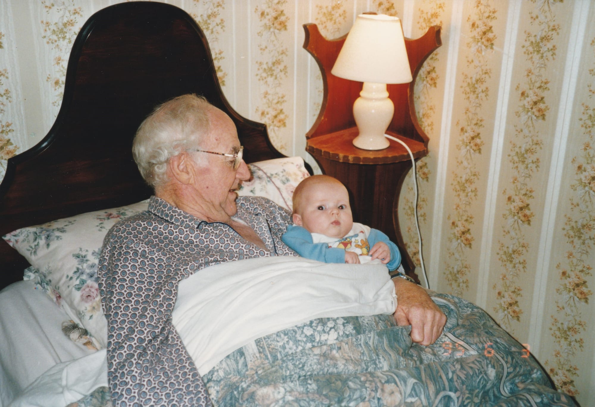 A 73 year old man, in formal button-up pajamas, tucked into his bed with a 6-month old baby in Winnie the Pooh pajamas in his arms. He is smiling at the baby, and the baby is looking at the camera with concern.